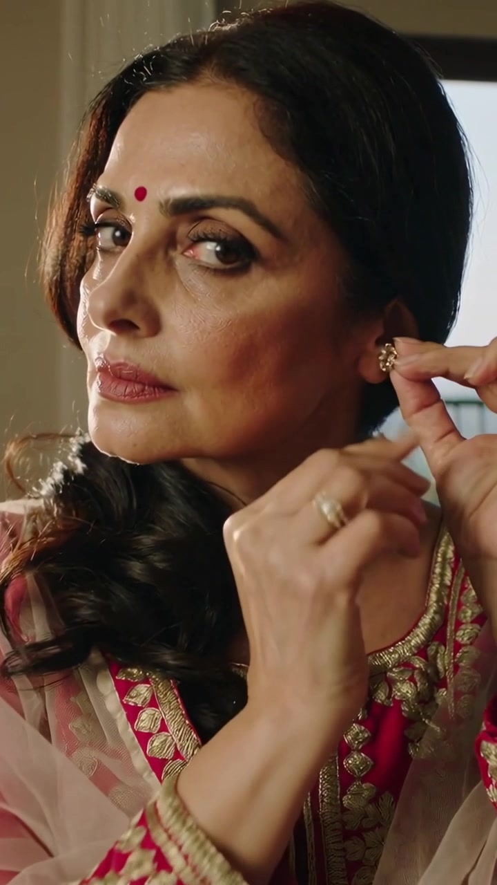 Vertical video: Indian woman putting on earring in red kurta by window at home, preparing for event