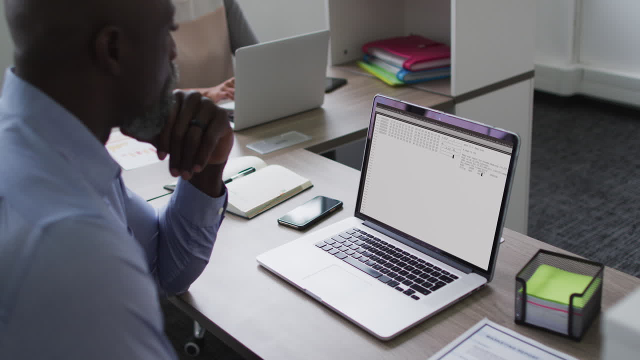 Afroamerikansk mand sidder ved skrivebord og ser på kodning og databehandling på laptopskærmen.