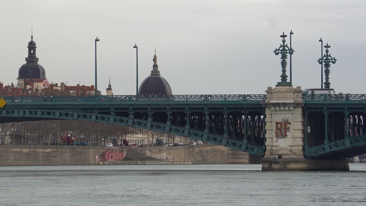 버스와 보행자 들 은 리옹 의 오래된 철 대학 다리 를 건너고 있다. 이 다리 는 아치 와 장식적 인 도자기 로 된 기둥 으로 장식 되어 있다.