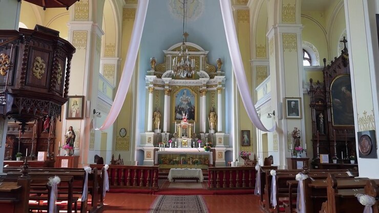 Splendida ripresa grandangolare di un interno di chiesa vuoto e ornato, inondato di luce solare, con dettagli intricati dell'altare, banchi di legno, lampadari e drappeggi nuziali bianchi, ripresa con telecamera stabile.