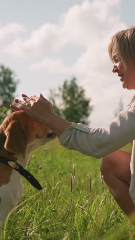 woman feeding dog in vast grassy field while squatting on sunny day, sharing affectionate bond with her dog gazing lovingly, surrounded by lush green nature and trees
