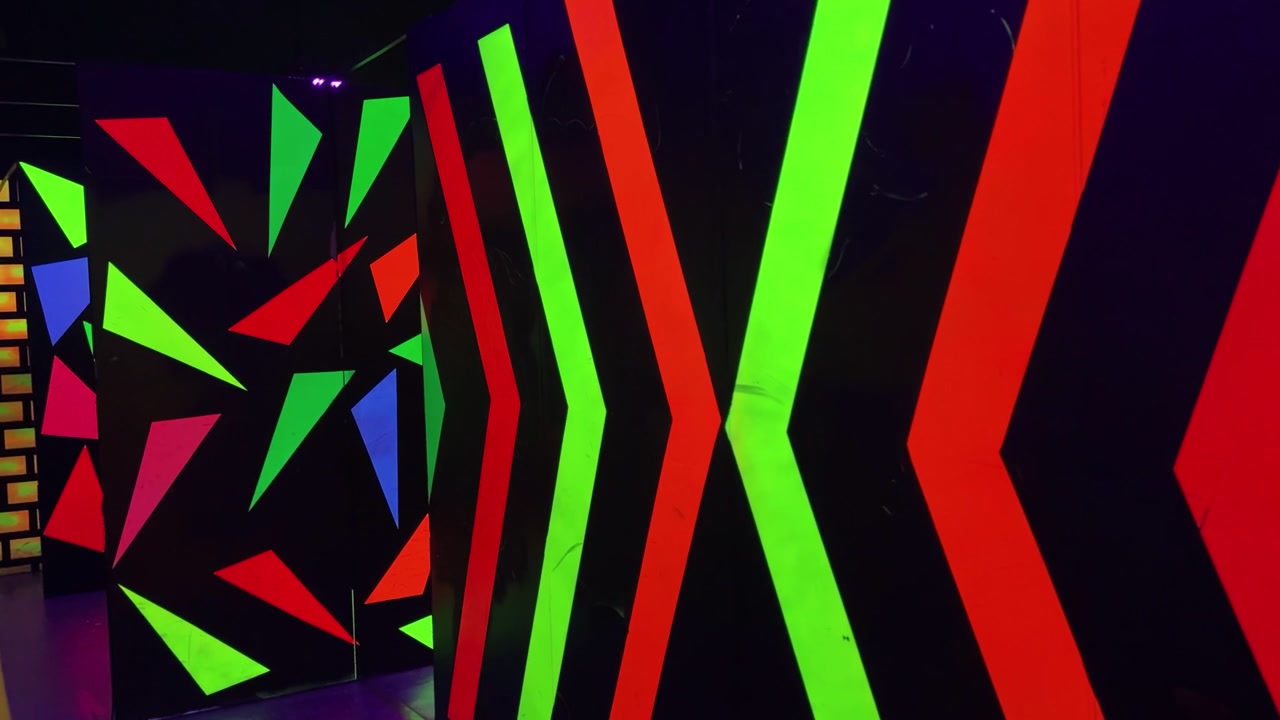 A static shot of an empty indoor entertainment center featuring dark maze walls illuminated by ultraviolet blacklight. Bright neon geometric shapes and arrows glow vibrantly in the laser tag arena.