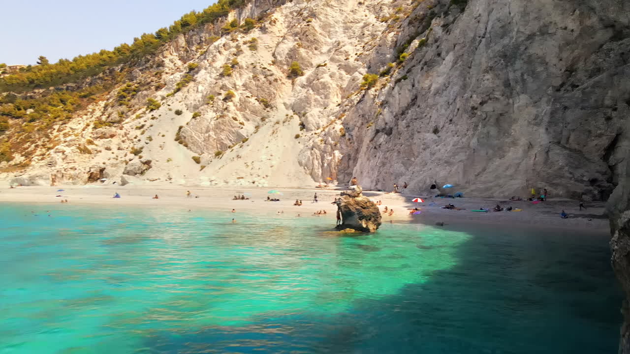 Aerial drone view of the Ionian Sea coast of Zakynthos, Greece. Rocky cliff, vegetation, beach with resting people, blue water. Slow motion