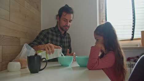 Happy-brunette-man-father-in-a-green-checkered-shirt-pours-milk-into-blue-plates-for-himself-and-his-daughter-brunette-girl-in-a-pink-dress-during-breakfast-at-the-table-in-the-kitchen