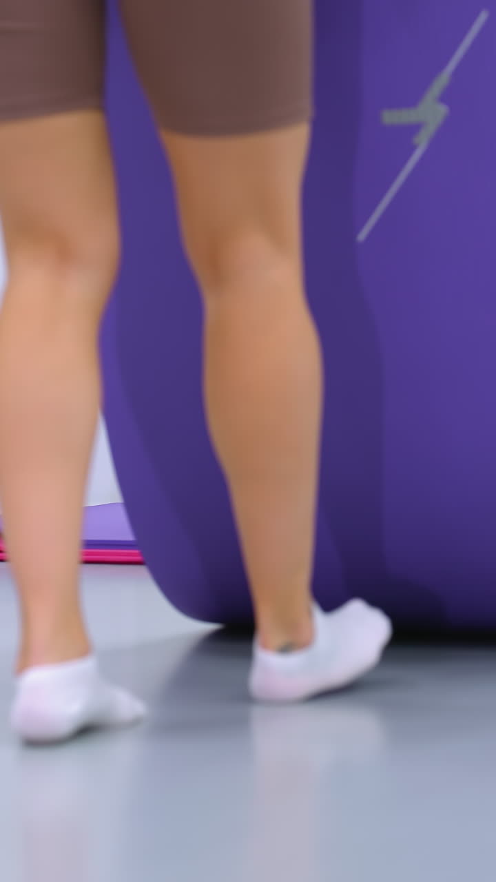 갈색 스포츠 의류를 입은 운동선수 여성이 피트니스 스튜디오에서 보라색 요가 매트를 들고, 거울은 그녀의 움직임을 반영하여 배경에 운동 매트와 장비가 있는 체육관 환경을 보여줍니다.