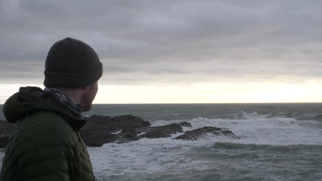 man wearing winter clothes overlooks stormy sea at sunset