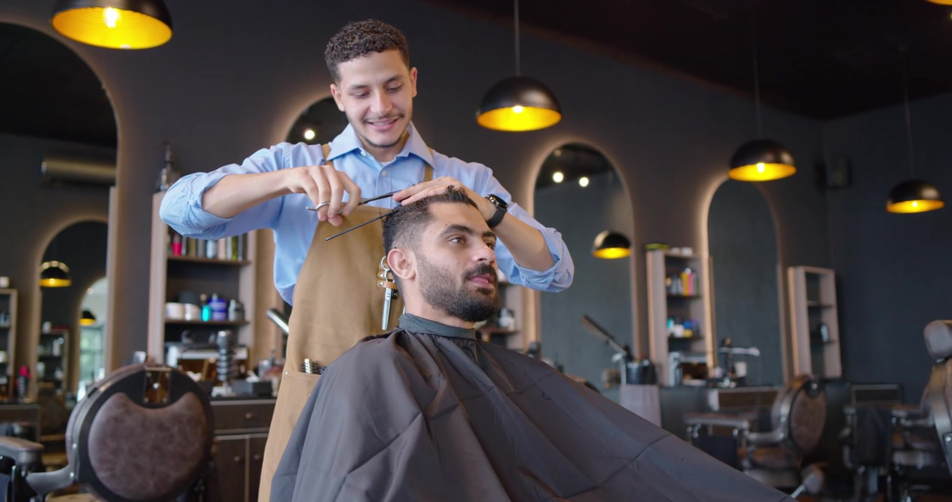 Barber og kunde, barber forbereder, reder og klipper hår med saks for at færdiggøre klipningen i salonen.