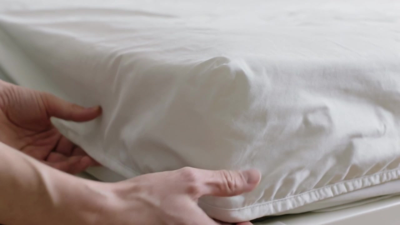 Lifting hands, pulling elastic edge down over mattress corner in bedroom to smooth fitted sheet