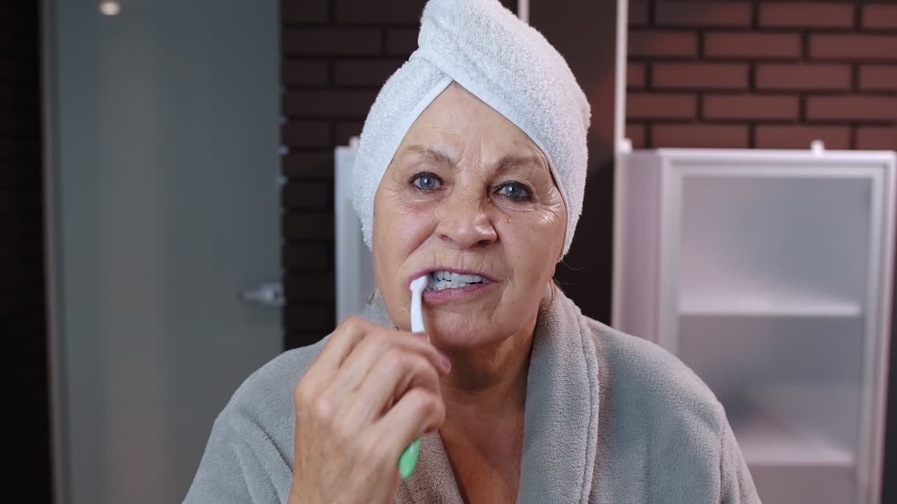 abuela mayor con toalla cepillándose los dientes y mirándose en un espejo después de la ducha en casa