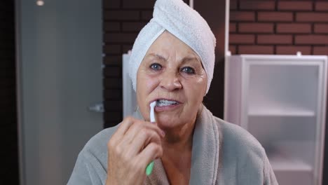 Abuela-Anciana-Con-Toalla-Cepillándose-Los-Dientes-Y-Mirándose-En-Un-Espejo-Después-De-La-Ducha-En-Casa