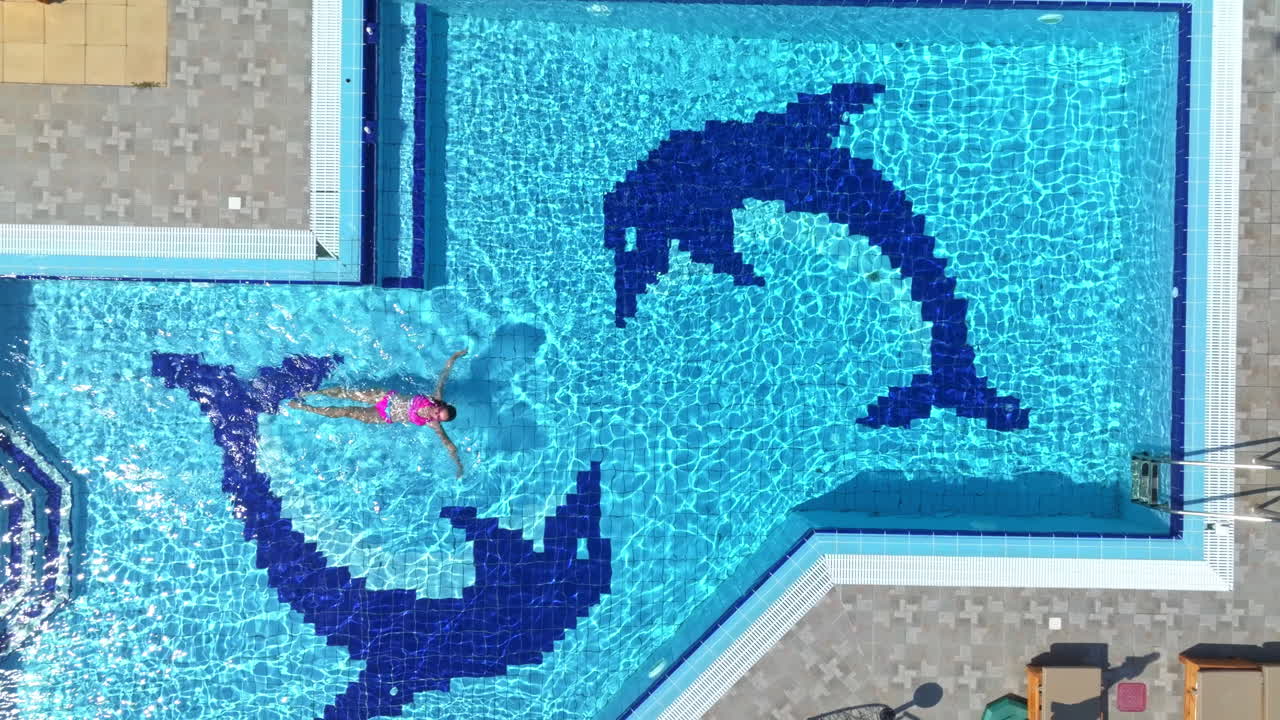 Birds eye drone shot above a woman swimming in a pool at a villa, sunny day
