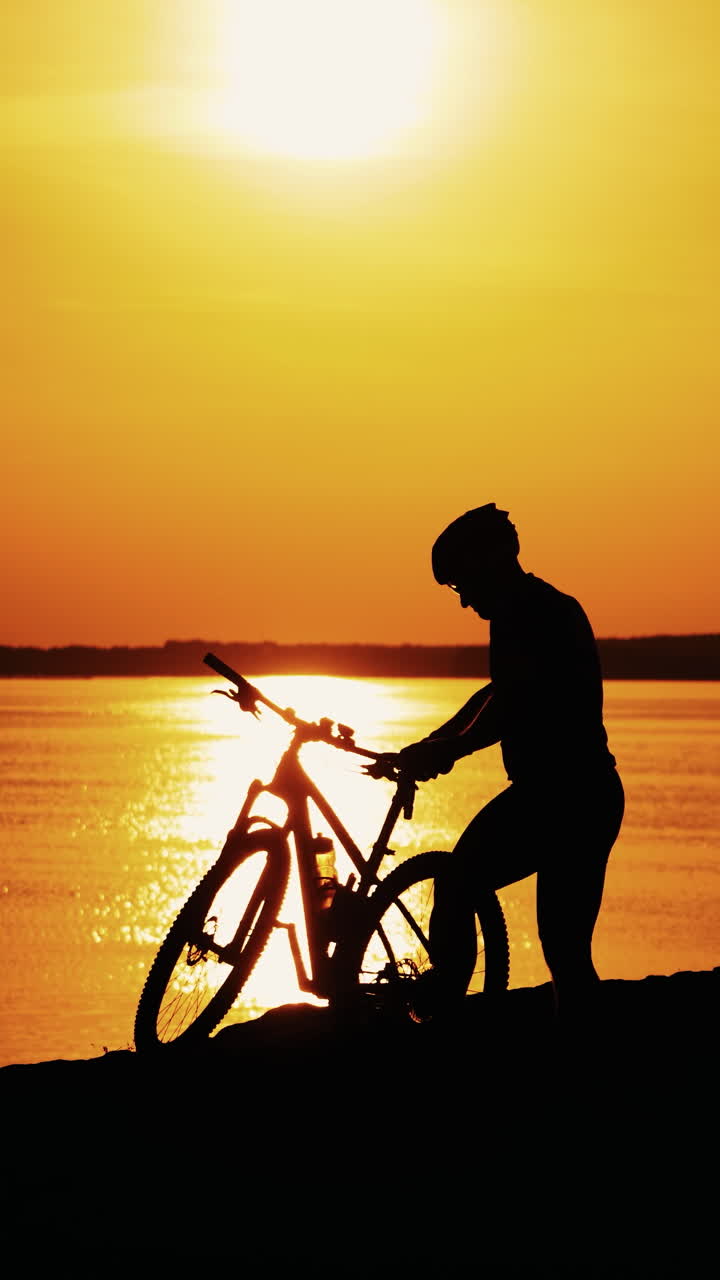 Professional male biker gets off the bike near the river at a beautiful sunset. Silhouette of a sporty cyclist in helmet on the evening water background at orange setting sun. Vertical video