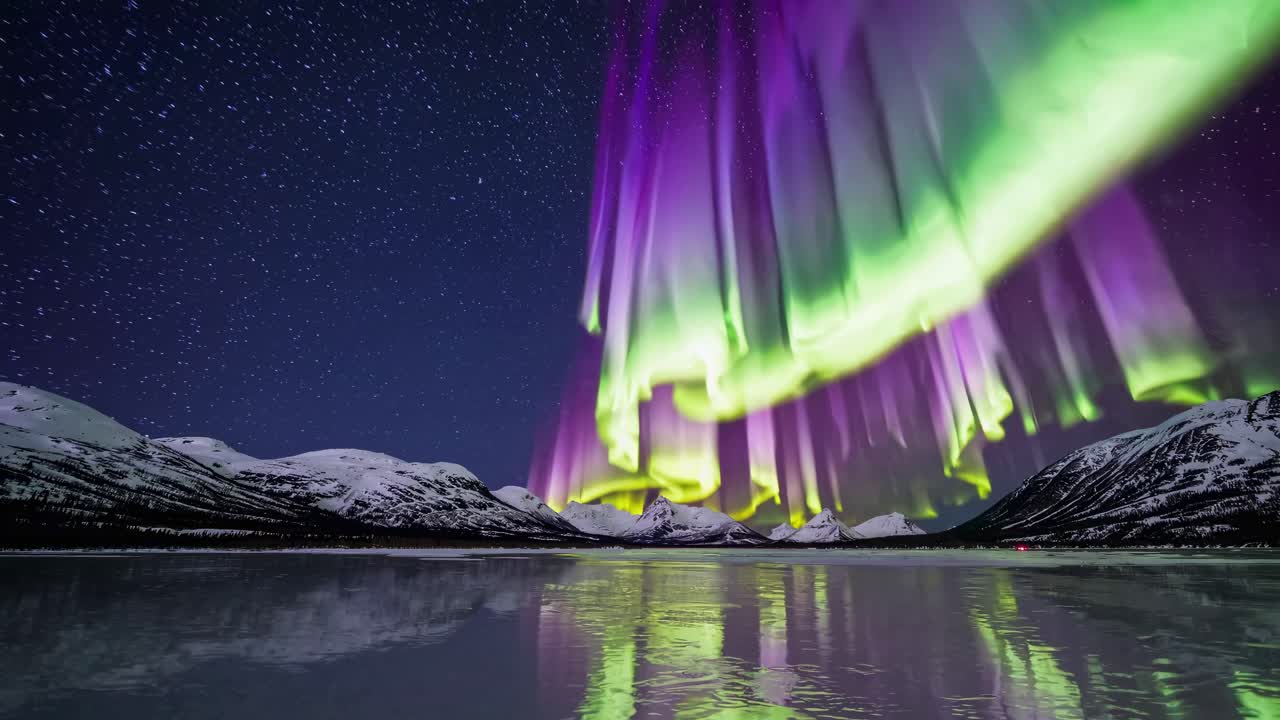 Wide-angle video capture of vibrant aurora borealis over snowy mountains, reflecting on a frozen