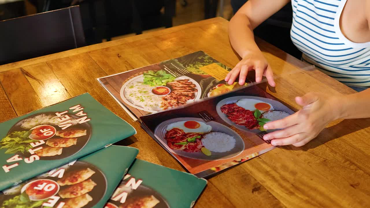 A person browses a vibrant Vietnamese food menu in a Bangkok restaurant, highlighting diverse dishes under warm lighting