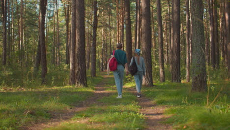 back view of two sisters walking along a forest trail surrounded by lush greenery, one sister wears a blue hair tie and carries a black backpack, while the other has a red backpack