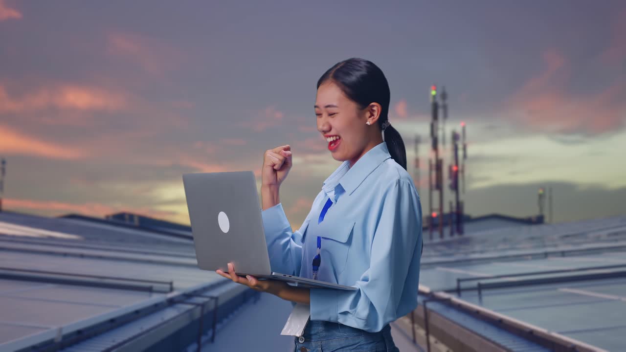 亞洲女性手持筆記型電腦的側面照，她正在檢查工業大廈天台太陽能板，在筆記型電腦上輸入後興奮地高舉拳頭歡呼。