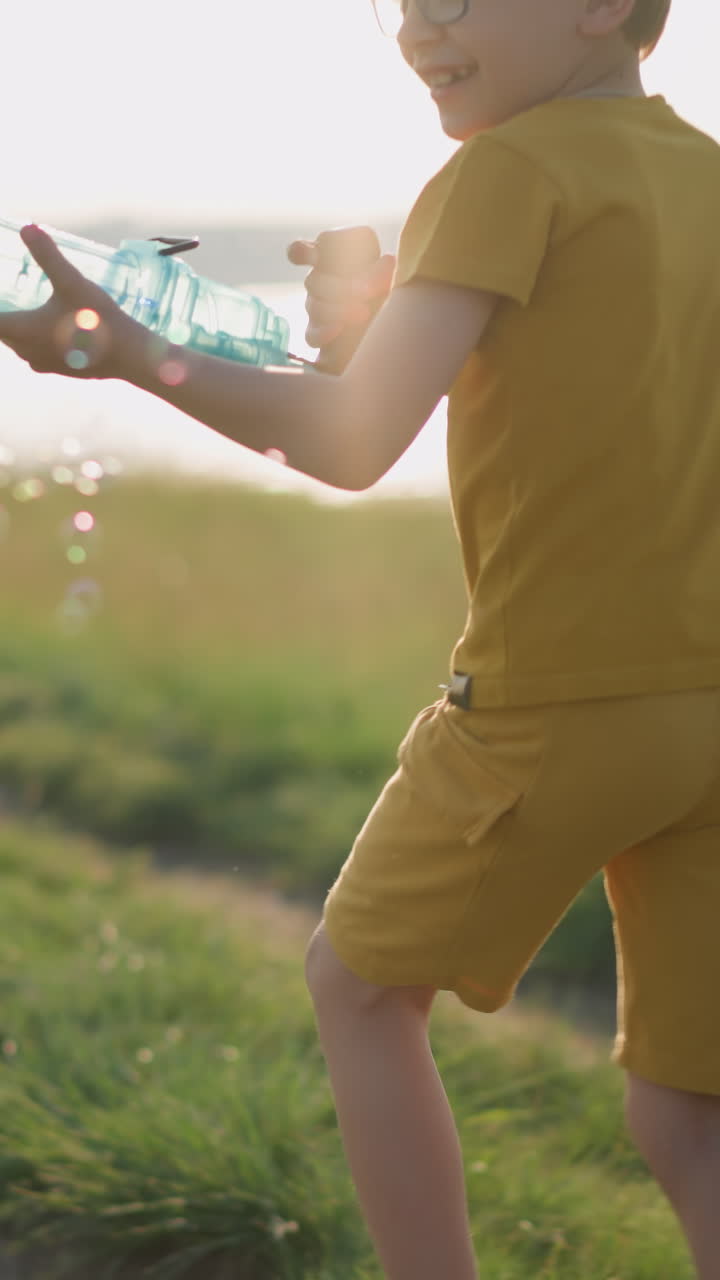 Young boy wearing a yellow shirt and shorts, with glasses, runs through a sunny grassy field while holding a plastic bubble gun that releases a stream of bubbles. The scene is set beside a serene lake