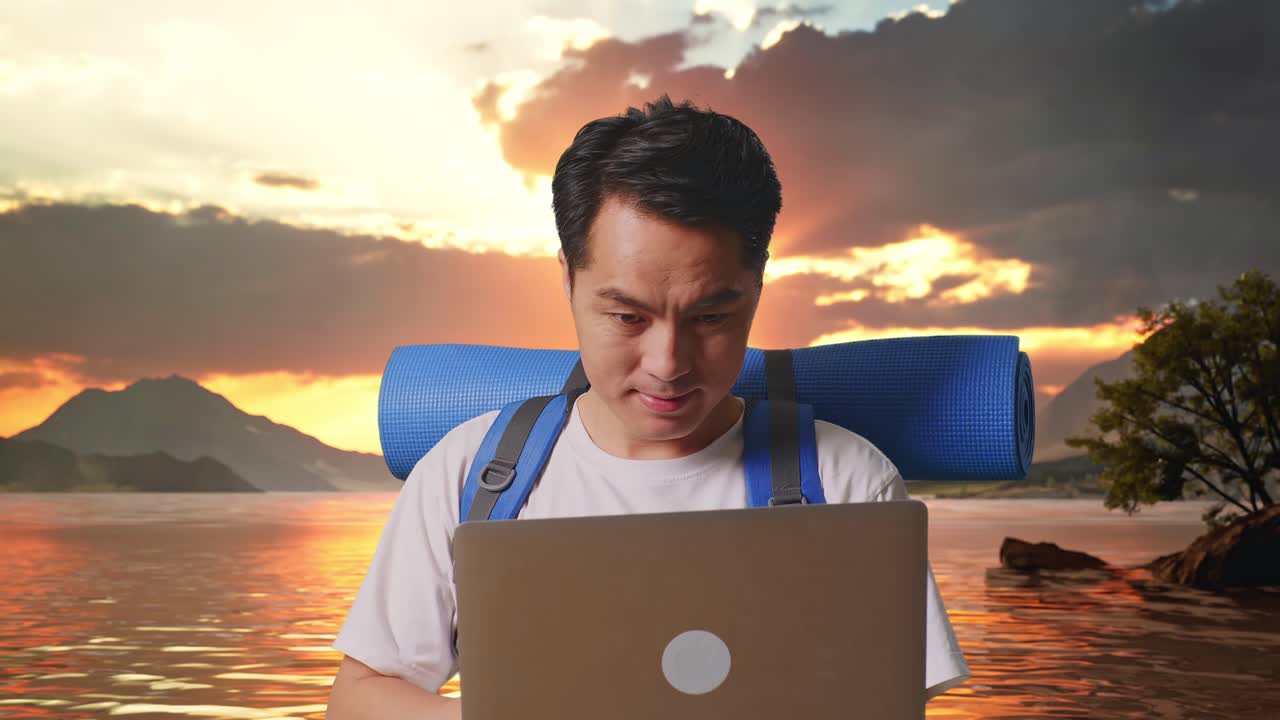Close Up Of Asian Male Hiker With Mountaineering Backpack Using A Laptop At A Lake