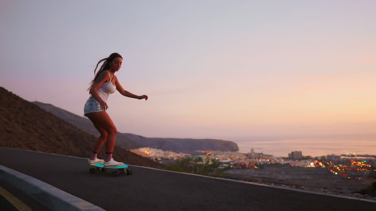 In slow motion, a breathtaking young skateboarder, dressed stylishly in shorts, rides her board along a mountain road at sunset, capturing the mountains' captivating view