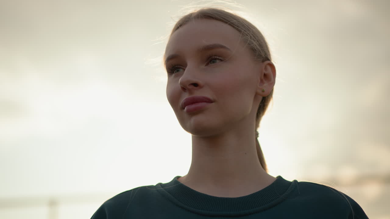 A lady in a green top with pink lips looks thoughtfully towards the horizon under a glowing sky, captured in a peaceful moment of contemplation, with sunlight glowing around her