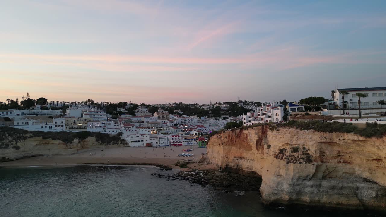 Drone che vola da destra a sinistra nella città di Carvoeiro, Algarve, Portogallo, al tramonto (ora d'oro), mostrando una spiaggia cittadina vuota e luci, ristoranti e bar in lontananza