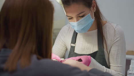 nail technician in white shirt, black apron, pink gloves and mask presents nail polish color samples to customer for selection while sitting at salon table under lamp during manicure session