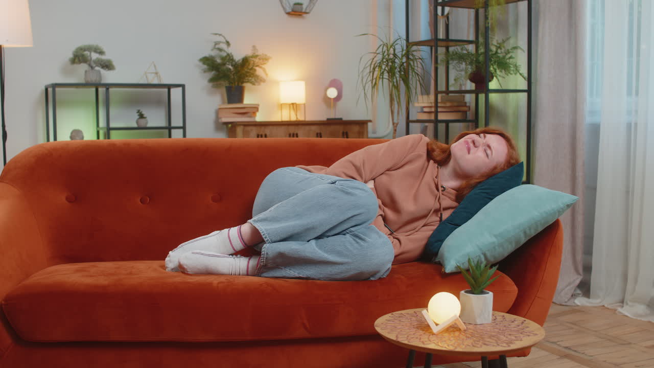Sick ill redhead woman suffering from period cramps painful stomach ache on sofa couch at home
