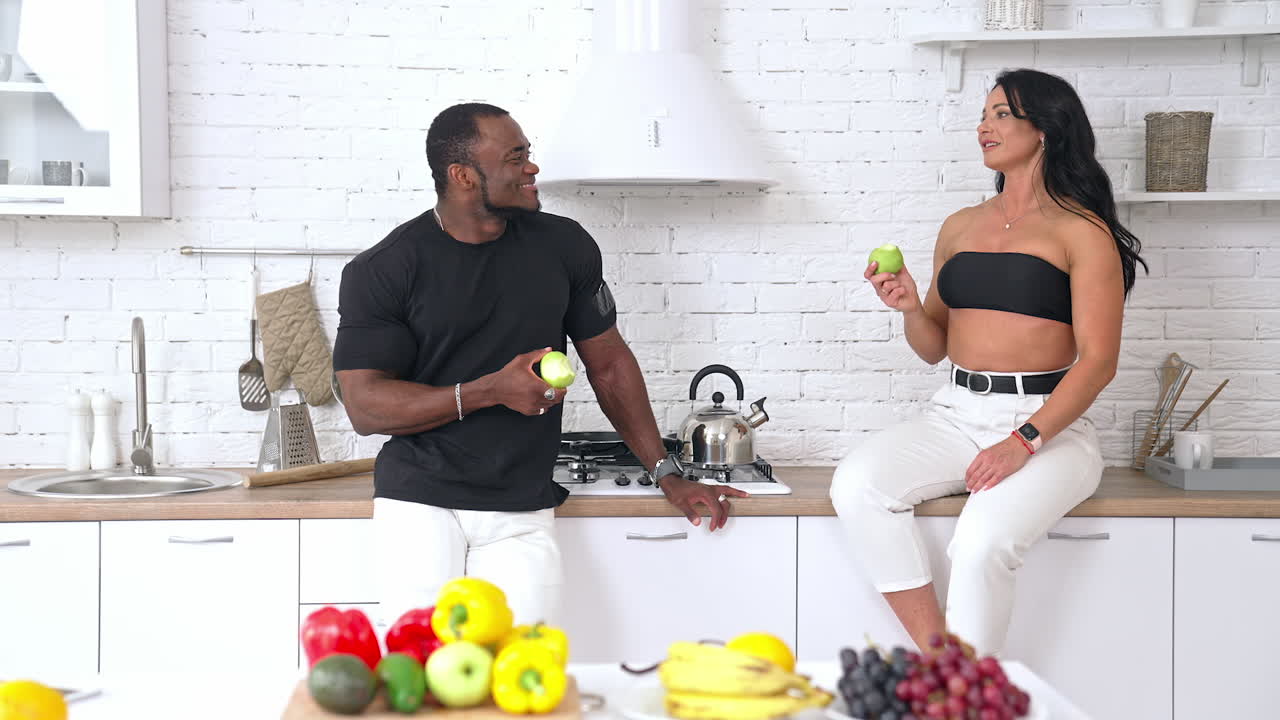 Sporty couple having a conversation at home. Caucasian woman sitting on the kitchen table and talking to afro man. Multi ethnic couple eating fresh apples. Healthy eating.