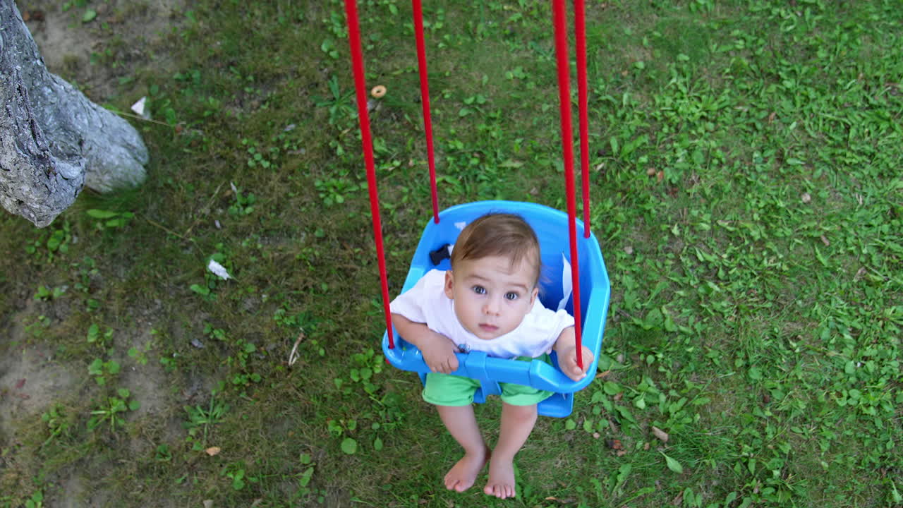 Curious small kid swaying in the swing. Baby looks up at camera. View from above. Green grass at backdrop.