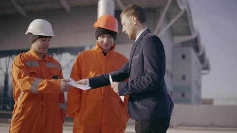 Manager-in-a-suit-giving-envelopes-with-money-to-workers-in-orange-uniform-and-helmets.-Successful-finish-of-the-project