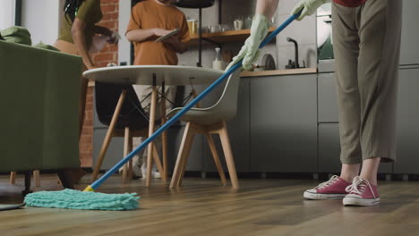 close up of an unrecognizable woman mopping the floor while her two roommates cleaning the kitchen