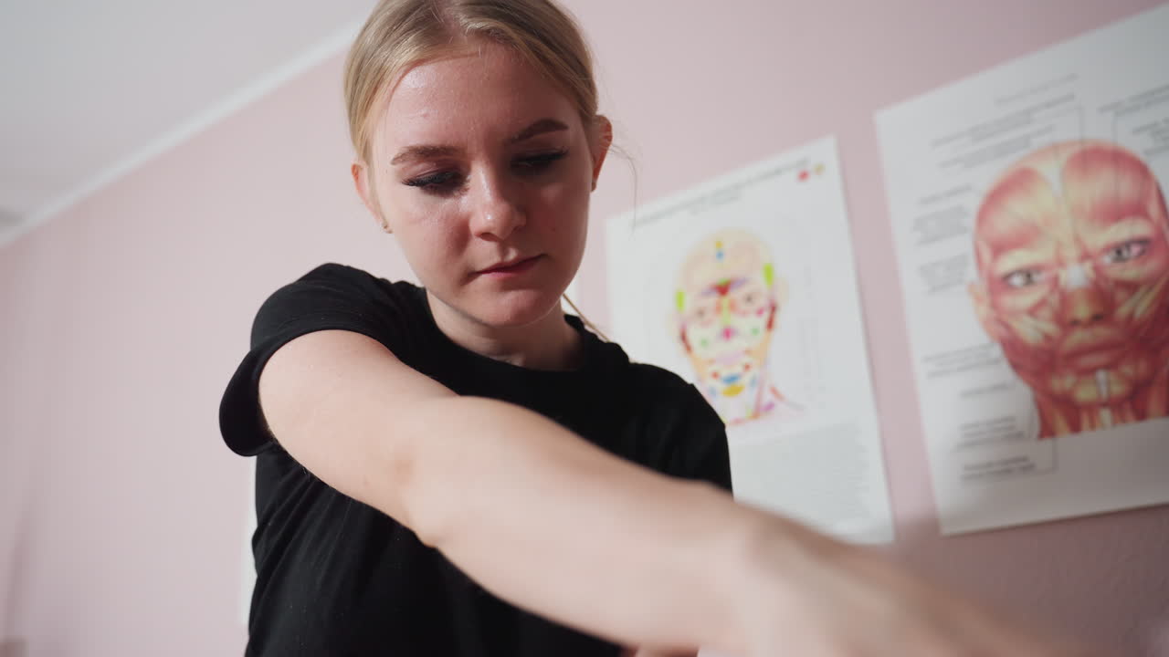 Massage therapist holding and stretching client foot during therapy session, focus on heel and sole with therapist face partially visible, anatomical muscle sketch on pink wall in background