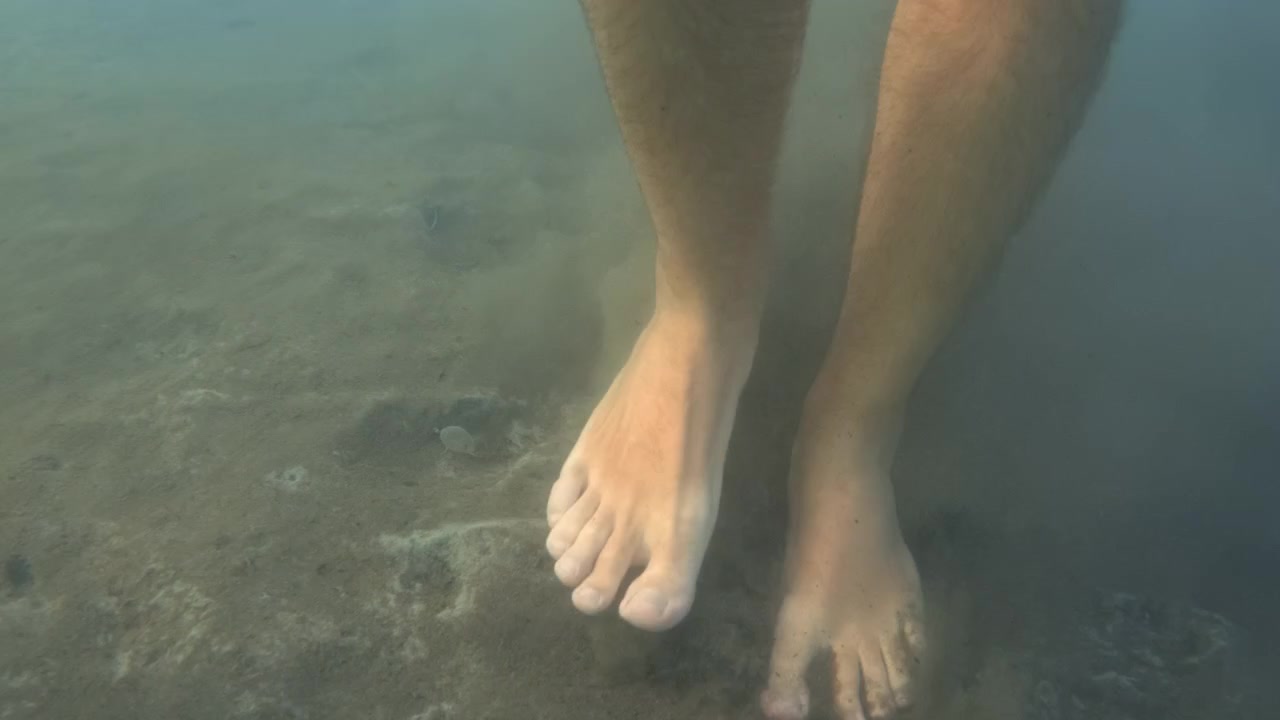 Gambe umane immerse in acqua blu sfocata. Che smuovono sabbia e limo sul fondale marino, creando un effetto torbido