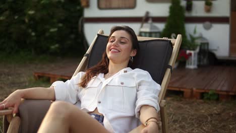 Pleased,-attractive-caucasian-woman-chilling-out-and-smiling-while-laying-on-lounger-outdoors.-Traveling,-rest,-vacation-concept.-Trailer,-wheels-house-on-the-blurred-background