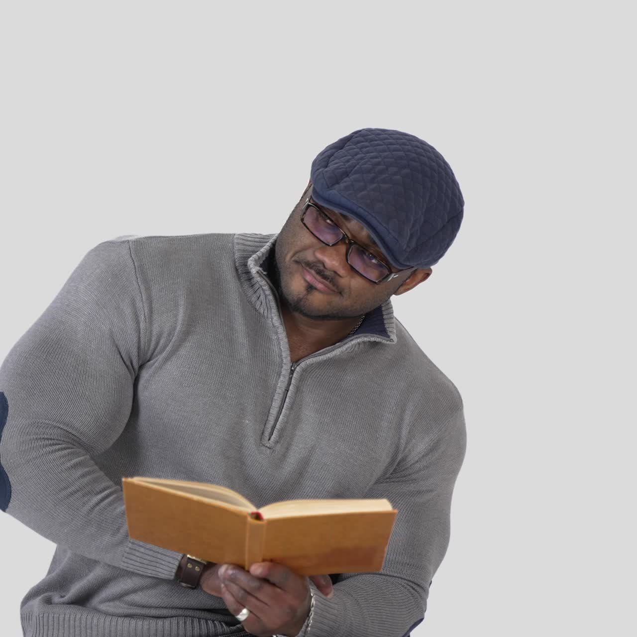 Serious african man reading book. Black skinned guy wearing glasses, cap and sweater sitting on chair with a book isolated on white