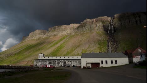 fotografía panorámica de la bahía de djúpavík con montañas, cascadas y antigua fábrica durante el cielo nublado gris al atardecer, islandia