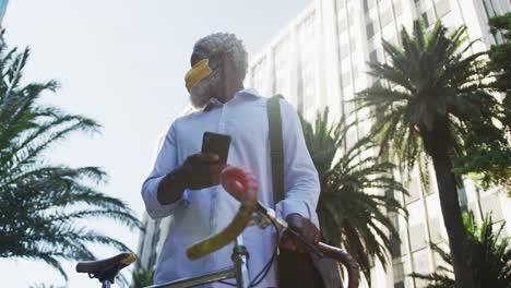 hombre mayor afroamericano con máscara facial con bicicleta usando un teléfono inteligente y mirando a su alrededor en co