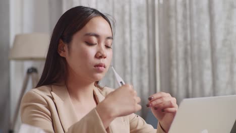 아픈 아시아 사업가 여성이 집에서 거실에서 노트북으로 일하는 동안 독감, 감기 및 신체 온도에 대한 온도 측정 테스트를 사용합니다.