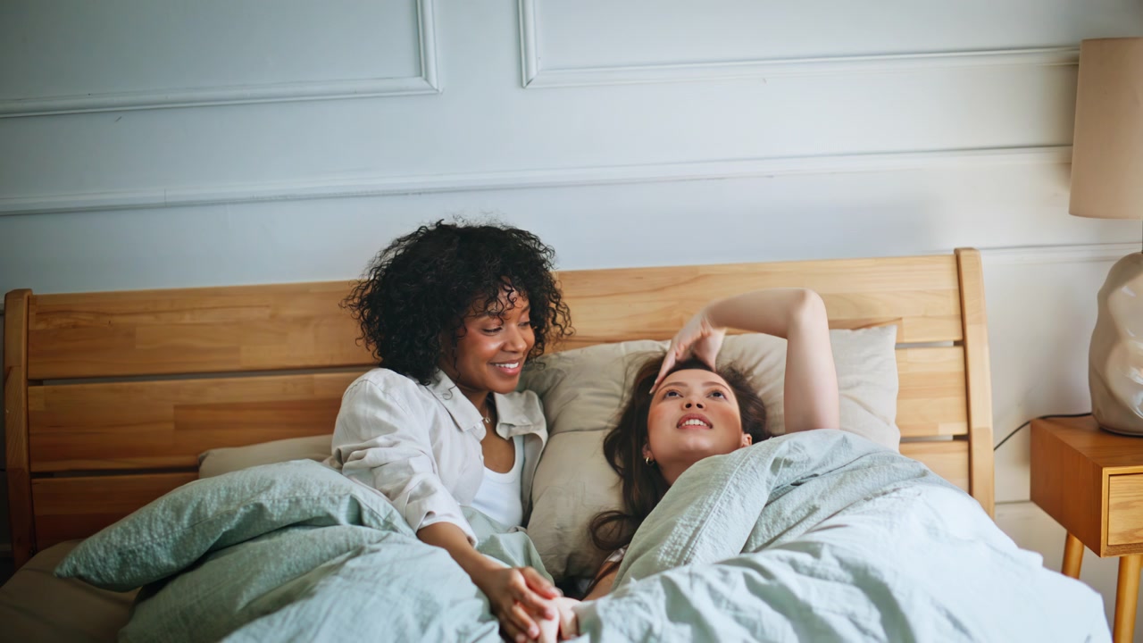 Lesbian pair waking bed holding hands. Woman stretching arm cozy bedroom.