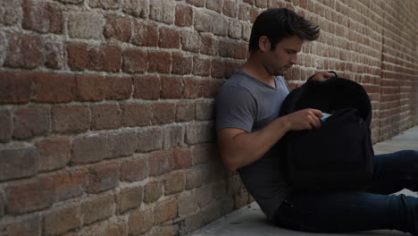 man sits by a brick wall searching through his backpack