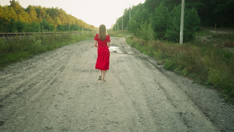 excited woman in red dress walking freely along countryside dirt road near railway track surrounded by green forest and soft evening light in peaceful natural outdoor setting