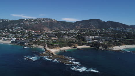 Panning-aerial-drone-shot-over-the-beautiful-blue-water-of-Laguna-Beach,-California-with-the-mountains-in-the-background