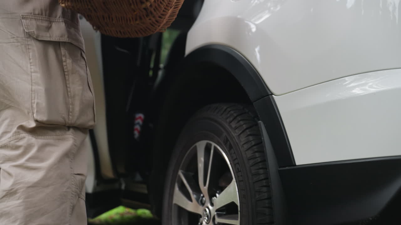 젊은 남자가 음식과 담요가 가득 담긴 피크닉 바구니를 들고 숲길을 걸어 차로 향하는 낮은 앵글의 시점이에요. 평화로운 가족 나들이 분위기를 보여주는 캐주얼한 여름날의 풍경입니다.