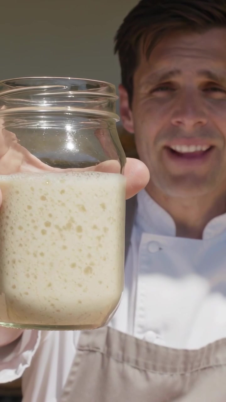 Video verticale: Chef in camice bianco e grembiule beige solleva un barattolo di vetro sulla soglia, mostrando il lievito madre.