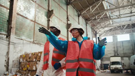 A-brunette-girl-in-safety-glasses-and-a-special-uniform-in-an-orange-vest-and-a-white-helmet-walks-with-her-employee-a-man-with-Black-skin-through-a-huge-waste-paper-processing-plant