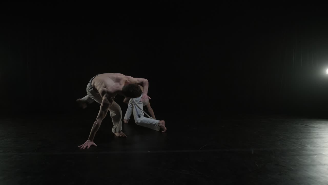 Group of a professional dancers are practicing capoeira in darkness against a spotlight on a black background of studio. Afro-Brazilian martial art that combine elements of dance.