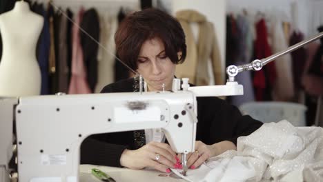 Retrato-De-Una-Costurera-Adulta-Trabaja-En-Una-Máquina-De-Coser,-Trabajando-En-Un-Vestido-Blanco-Hecho-A-Mano.-Ropa-De-Moda-Y-Maniquí-Blanco-En-El-Fondo