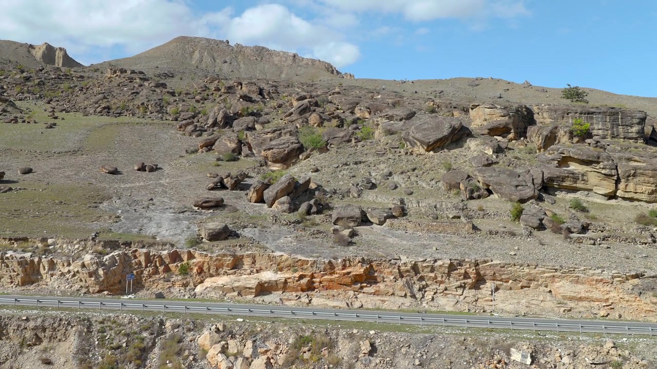 Ampia vista stradale attraverso una ripida collina rocciosa e accidentata con scogliere a strati, massi sparsi, vegetazione rada e un guardrail basso sotto un cielo azzurro brillante con nuvole soffici, che mostra ruvide texture terrose