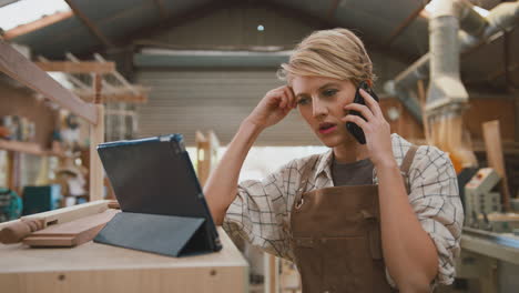 frustrated female carpenter with digital tablet working in furniture workshop making phone call