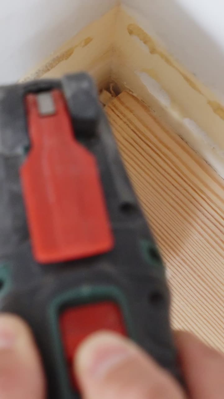 person using a triangular orbital sander to smooth the corner of a wooden floor near a wall, focusing on precision during home improvement work, vertical close-up shot